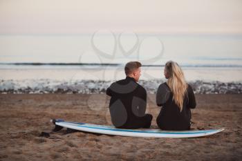 Rear View Of Thoughtful Couple Wearing Wetsuits On Surfing Staycation Looking Out To  Sea At Waves