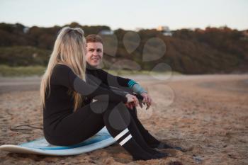 Couple Wearing Wetsuits On Surfing Staycation Sitting On Surfboard Looking Out To  Sea At Waves