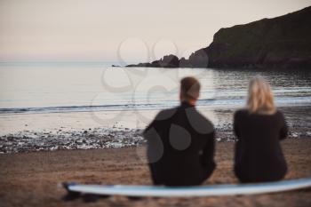Defocused Shot Of Couple In Wetsuits On Surfing Staycation Sitting On Surfboard Looking Out To  Sea