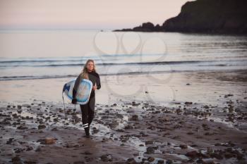 Woman Wearing Wetsuit Carrying Surfboard As She Walks Out Of Sea