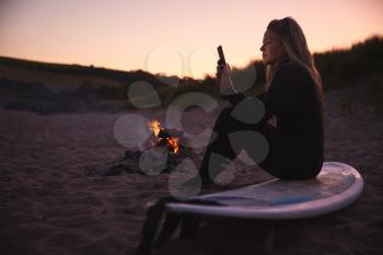 Woman Sitting On Surfboard By Camp Fire On Beach Using Mobile Phone As Sun Sets Behind Her