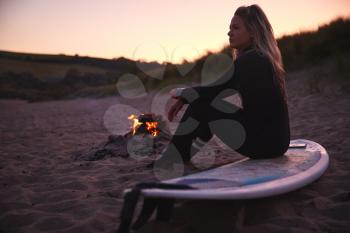 Woman Sitting On Surfboard By Camp Fire On Beach As Sun Sets Behind Her