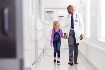 Male Paediatric Doctor Walking Along Hospital Corridor Holding Hands With Young Girl Patient