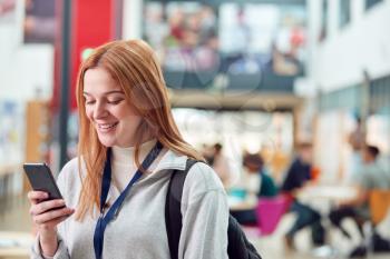 Smiling Female College Student Checking Mobile Phone In Busy Communal Campus Building