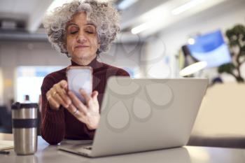 Mature Businesswoman With Travel Mug Working On Laptop In Modern Office Using Mobile Phone