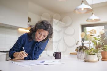 Mature Woman Reviewing And Signing Domestic Finances And Investment Paperwork In Kitchen At Home