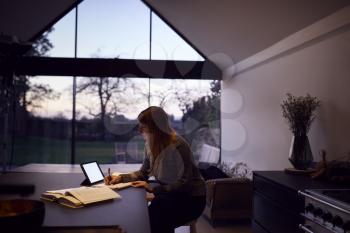 Evening Shot Of Woman In Kitchen Working Or Studying From Home Using Digital Tablet