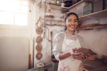 Portrait Of Female Potter Wearing Apron Holding Lump Of Clay In Ceramics Studio