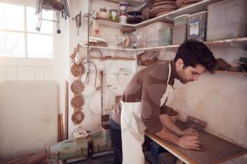 Male Potter Wearing Apron Rolling Out Lump Of Clay In Ceramics Studio