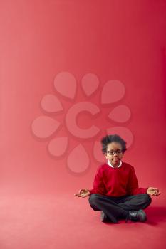 Male Elementary School Pupil Wearing Uniform Sitting And Meditating Against Red Studio Background