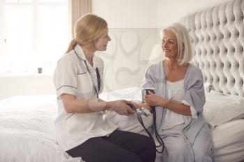 Female Doctor Making Home Visit To Senior Woman In Bedroom For Blood Pressure Check