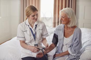 Female Doctor Making Home Visit To Senior Woman In Bedroom For Blood Pressure Check