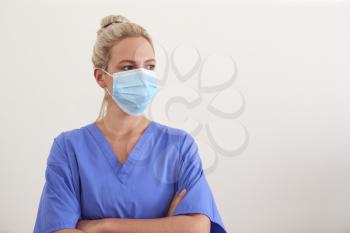 Studio Portrait Of Female Nurse Wearing Scrubs And PPE Face Mask Against White Background