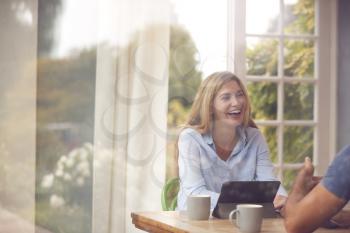 Couple With Digital Tablet Sitting At Table Working From Home Viewd Through Window