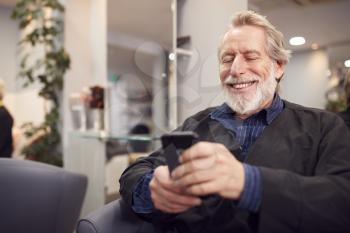 Senior Man Waiting To Have Hair Cut In Hairdressing Salon Looking At Mobile Phone