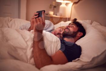 Couple With Man Lying In Bed At Night Looking At Mobile Phone Screen