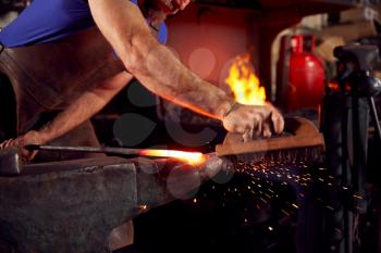 Close Up Of Male Blacksmith Using Wire Brush On Metalwork Resting On Anvil With Sparks