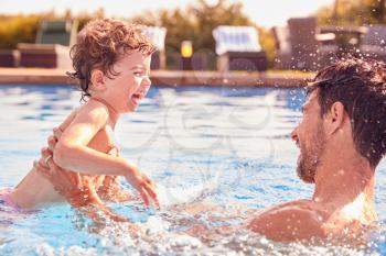Father With Young Son Having Fun On Summer Vacation Splashing In Outdoor Swimming Pool