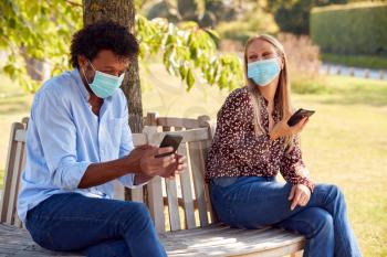 Couple Wearing Masks Meeting In Outdoor Park During Health Pandemic Looking At Mobile Phones