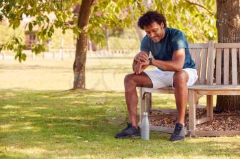 Man Wearing Fitness Clothing Sitting On Seat Under Tree Checking Activity Monitor On Smartwatch