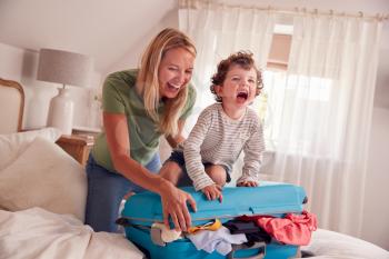 Mother And Son Packing For Vacation With Boy Standing On Full Suitcase To Close
