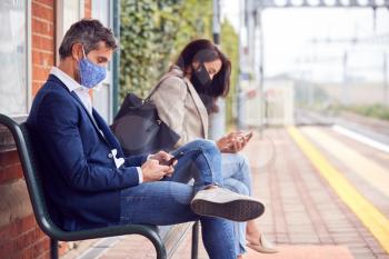 Business Commuters Sitting On Railway Platform With Mobile Phones Wearing Face Masks During Pandemic