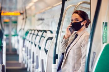 Businesswoman In Train Carriage Talking On Mobile Phone Wearing PPE Face Masks During Pandemic