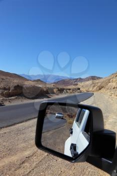 The asphalted road to Death Valley. On a roadside there is a white car. In its mirror of the distant review the road and other white car is reflected