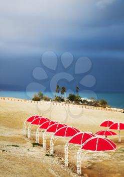 Red beach umbrellas on deserted coast of the Dead Sea in thunder-storm 