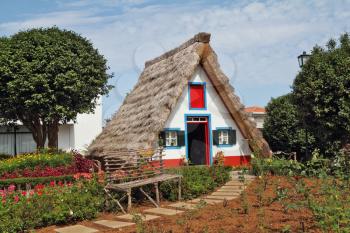 Classical triangular small house of the first settlers on island Madiera. A bright red door, a straw triangular roof, a small front garden and an accurate stone path
