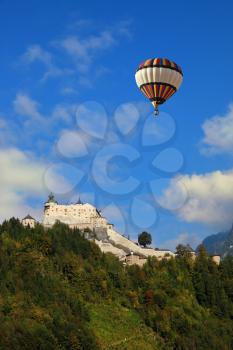 The castle is situated on top of the mountain and surrounded by dense forest.  Majestic medieval Palase Hohenwerfen.  Over the castle and the mountains in the clouds flying a giant balloon
