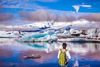 The woman - tourist with a green backpack watching the moving ice. Sunrise in the Ice Lagoon. Ice floes are reflected in the smooth water surface. The concept of extreme northern tourism