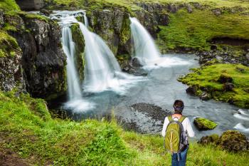 Elderly woman -  traveler with a large backpack admiring the powerful jets of the waterfall Kirkjoufellfoss. Summer in Iceland. Concept of exotic and extreme tourism 
