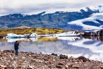 The elderly woman photographs the lagoon. The concept of extreme northern tourism. Ice floes are reflected in the smooth water surface of Ice Lagoon Jokulsarlon, Iceland