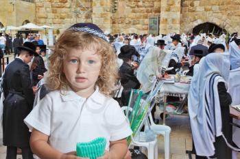 Adorable little boy with long blond curls and blue eyes in knitted skullcap. He stands at the main Jewish shrine - Western Wall of Temple. The Jewish holiday of Sukkot