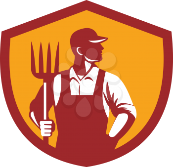 Illustration of organic farmer holding pitchfork looking to the side with one hand in pocket viewed from front  set inside shield crest on isolated background done in retro style. 