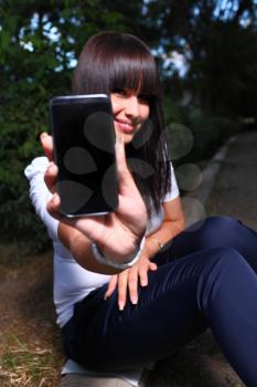 Smiling teenage girl shows cell phone, against green of summer park in the dark vertical focus on phone