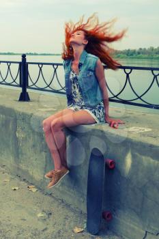 Young women sitting with her skate board on concrete street parapet with her red hair flying in the wind