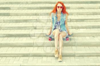 Beautiful redhead young woman sitting over a skateboard on street stairs as background