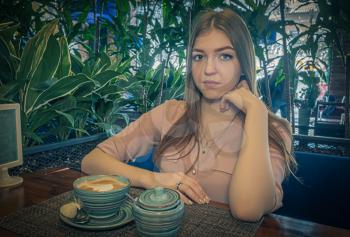 Nice lady sitting in cafeteria with milk coffee with froth on top served in front on her with sugar bowl aside. She is looking at camera and touching her chin.