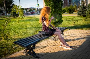 Ginger haired lady is sitting on park bench in trendy summer dress with small black handbag aside.
