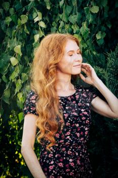 Portrait of a serious beautiful redhead women closed eyes touching her face and looks relaxed and enjoying the fair weather in garden.