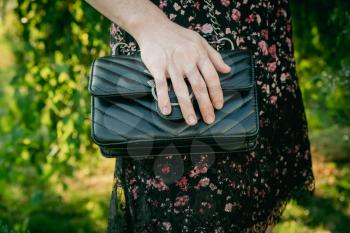 Stylish lady in summer dress walking alone in park with black leaser handbag. Summertime in the park: Young woman in black dress with floral ornament posing with handbag with her palm on