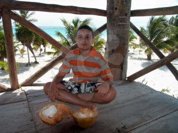A boy with coconuts in the hut built on the caribbean beach, with the sunscreen covered face.