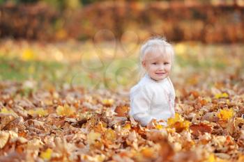 Little 3-4-year-old blonde girl sits on fallen leaves and smiles while walking in the autumn park