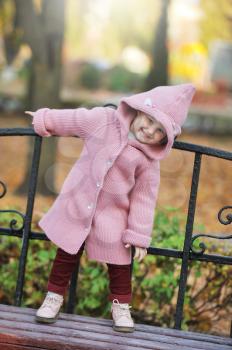 A little blonde girl 3-4 years old in a knitted coat with a hood stands on a bench and smiles against the background of an autumn Park during a walk