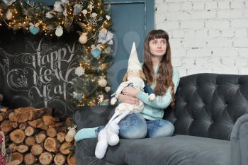 Young girl in jeans sits on a sofa with a soft toy dwarf on the background of Christmas decor and chalk inscription happy new year