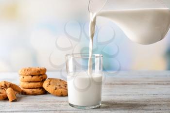 Milk pouring from jug into glass with cookies on wooden table�