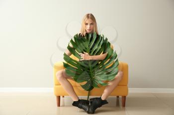 Beautiful woman with green tropical leaf sitting in armchair against white wall�