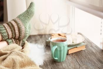 Cup of hot tea with legs of young woman on wooden table near window�
